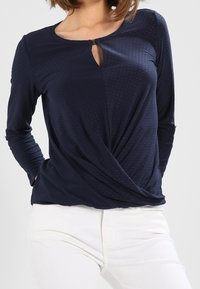 Kvinna som bär en marinblå långärmad topp med en liten nyckelhålsutskärning och snurrad framsida, i kombination med vita byxor.