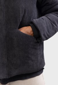 Chaqueta de pana negra con textura acanalada, que cuenta con bolsillos laterales y puños acanalados. Ajuste entallado con un forro suave y ligero.