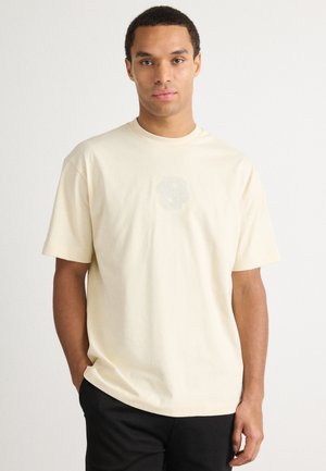 T-shirt in cotone crema con collo rotondo, maniche corte e logo "8" grigio chiaro sul petto. Il tessuto ha una consistenza morbida.