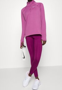 Personne portant un haut de sport à manches longues rose et des leggings violets, tenant une bouteille d'eau blanche avec le logo Nike noir, portant des baskets blanches.