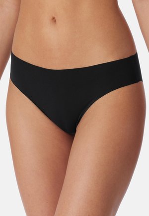 Frau trägt schlichte schwarze nahtlose Bikini-Unterwäsche und zeigt Bauch und Oberschenkel vor neutralem Hintergrund.