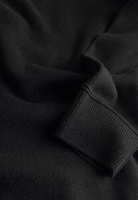 Tessuto nero lavorato a maglia con una texture morbida, caratterizzato da polsini a coste. Il materiale ha una finitura liscia e una caduta delicata.