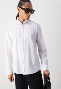 Camisa blanca de botones en tejido ligero con un acabado texturizado, con cuello y mangas largas, combinada con pantalones negros y gafas de sol.
