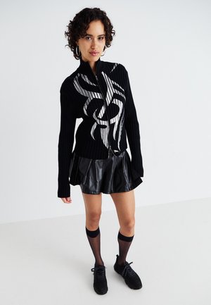 Svart ribbestrikket jakke med glidelås som har et sølvfarget abstrakt design, sammen med et svart faux skinn plissert skjørt, svarte sneakers og knelengde sokker.