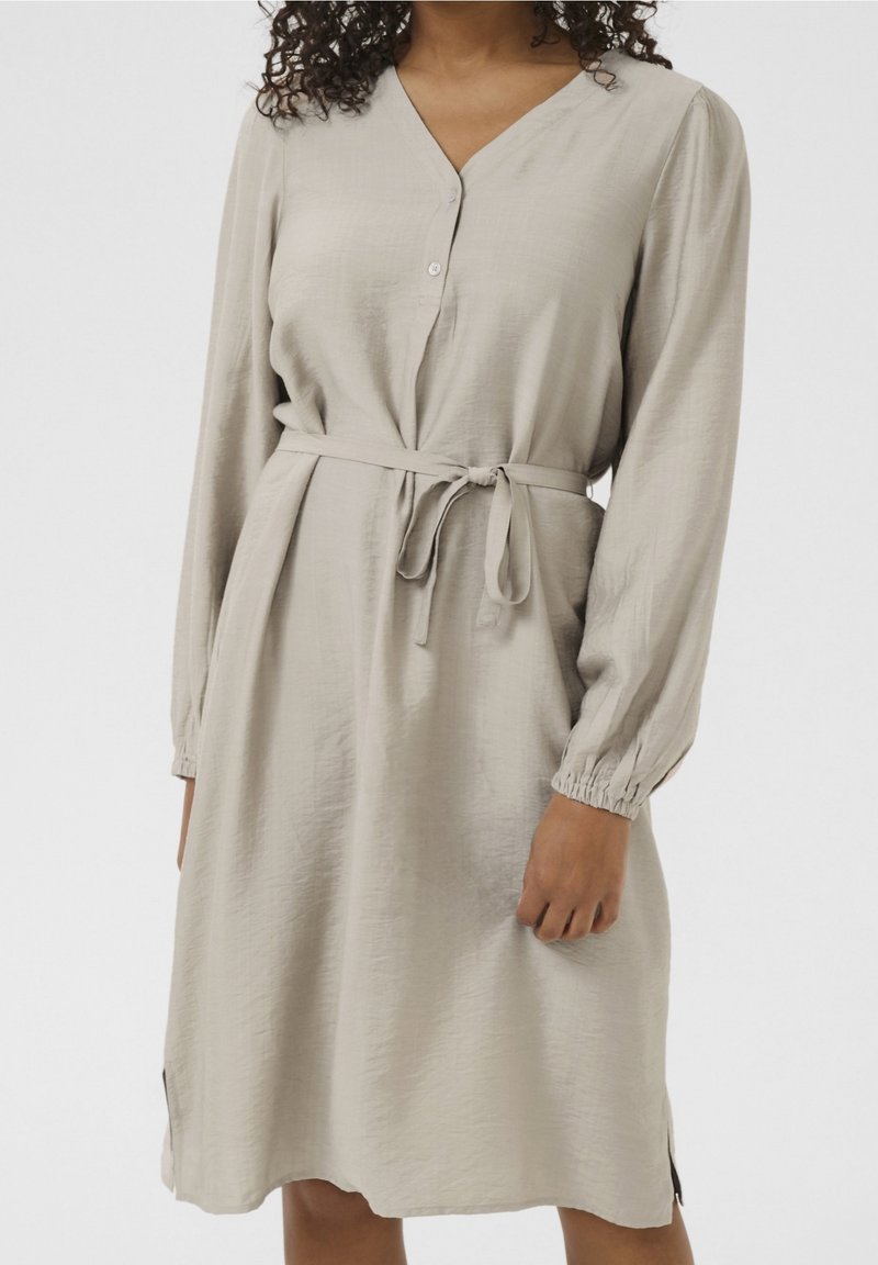 Femme portant une robe longue beige clair à manches longues avec un col en V, deux boutons et une ceinture en tissu nouée à la taille.