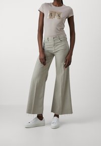Camiseta beige claro con logo A/X de lentejuelas, combinada con vaqueros de pierna ancha en gris claro con dobladillo deshilachado y zapatillas blancas.