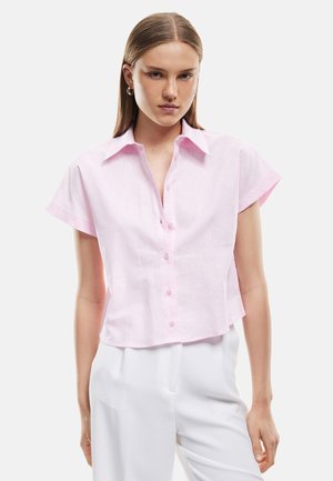 Kobieta z prostymi włosami nosi jasnoróżową krótką koszulę zapinaną na guziki i białe spodnie, stoi na prostym tle.