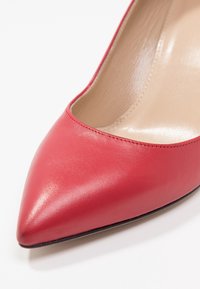 Gros plan sur une chaussure à talons aiguilles pour femmes en cuir rouge, à bout pointu, avec une doublure intérieure beige sur fond blanc.