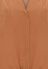Rostfarbene Kurzarmbluse mit glatter Textur, zwei vorderen Taschen und einer durchgehenden Knopfleiste mit passenden Knöpfen.