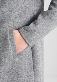 Grå textil som är mjuk i texturen, med en sidob Pocket. Designen inkluderar en rak skärning och långa ärmar.