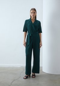 Groene plissé jumpsuit met een wikkelontwerp, korte mouwen, een riem en brede pijpen. Gemaakt met zwarte hoge hakken. Glad textuur.