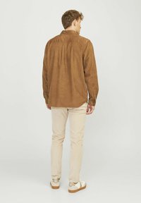 Chemise marron à manches longues avec un empiècement au dos et des poignets à boutons, associée à un pantalon beige clair et des baskets blanches. Texture de tissu lisse.