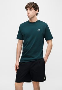 Junger Mann trägt ein dunkelgrünes New Balance T-Shirt und schwarze Shorts, steht mit einer Hand in der Tasche vor neutralem Hintergrund.