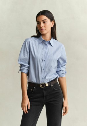 Femme aux longs cheveux foncés portant une chemise à rayures bleues et blanches à boutons avec les manches retroussées et un jean noir avec une ceinture.