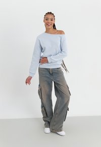 Lyseblå av- skuldre genser, sammen med overdimensjonerte beige cargo bukser med sidelommer, og hvite joggesko.