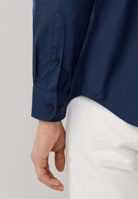 Granatowa koszula zapinana na guziki o teksturowanym wykończeniu, z dopasowanym mankietem z dwoma czarnymi guzikami oraz detalem wzdłuż bocznego szwu.