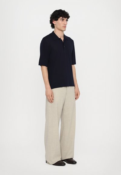 Polo shirt marine à manches courtes avec col, associé à un pantalon large beige clair et des chaussures foncées. Design et couleurs simples et classiques.