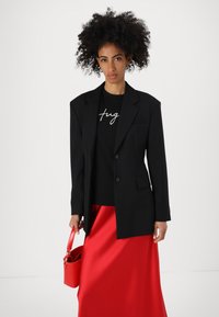 Black blazer with two buttons, worn over a black shirt with white text. Paired with a red satin skirt and a small red handbag.