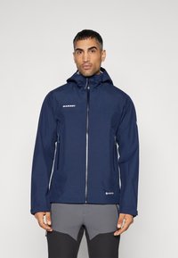 Marinblå vattentät jacka med huva, framdragkedja, två sidofickor och Gore-Tex-logga. Funktionen inkluderar en slät yta och hållbart material.