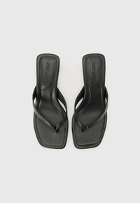 Des sandales en plastique noir avec une semelle plate, dotées de fines lanières et d'un design à bout carré. Fabriquées en matériau synthétique lisse avec un logo en relief.