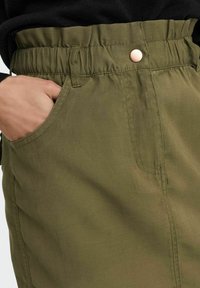 Jupe vert olive avec ceinture élastique et un seul bouton pression. Comprend des poches latérales et une texture douce et lisse.