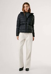 Melna polsterēta jaka ar kapuci, ar rāvējslēdzēja aizdari un šūtu rakstu. Valkāta virs pelēka džempera un kopā ar baltām biksēm.