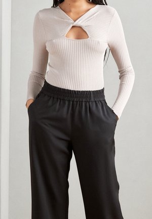 Femme portant un haut beige côtelé à manches longues avec un col découpé torsadé, associé à un pantalon noir taille haute avec taille élastique.