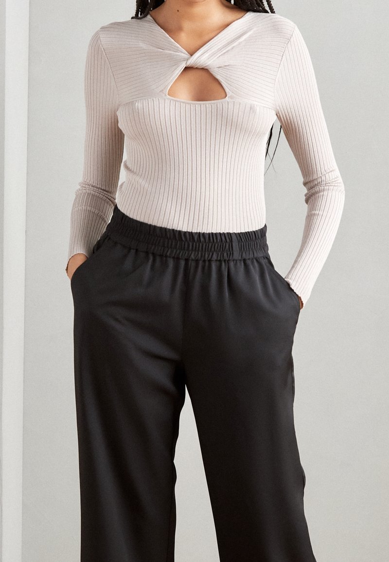 Femme portant un haut beige côtelé à manches longues avec un col découpé torsadé, associé à un pantalon noir taille haute avec taille élastique.