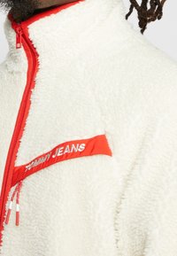 Giacca in pile color avorio con cerniera e bordi rossi. Presenta una toppa rossa con "TOMMY JEANS" ricamato in argento. Texture morbida.