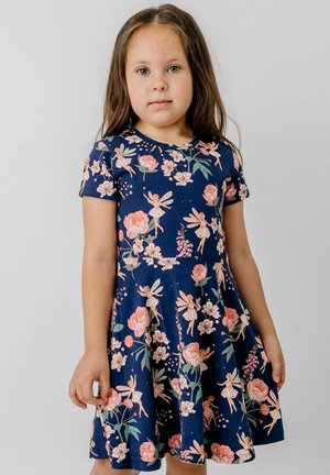 Junges Mädchen mit langen braunen Haaren trägt ein marineblaues Kleid mit pinkfarbenen Blumen und Feen, steht vor einem einfarbigen hellgrauen Hintergrund.