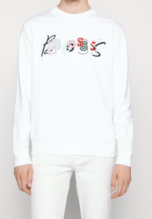 Person trägt ein weißes Langarmshirt mit bunten sich überlappenden Buchstaben, die "BOSS" über der Brust buchstabieren.