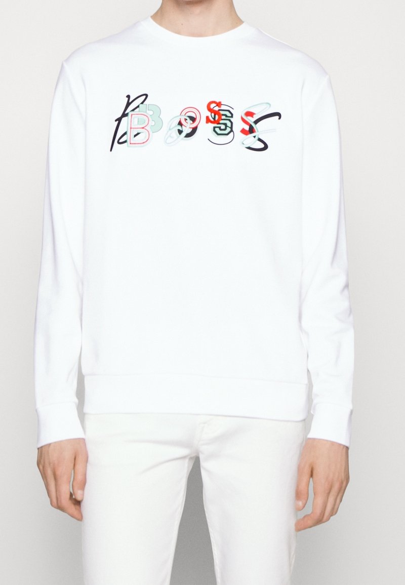 Personne portant une chemise blanche à manches longues avec des lettres colorées se chevauchant formant le mot "BOSS" sur la poitrine.