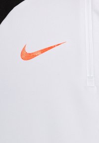 Top atletico bianco con design a yoke nero. Logo Nike arancione ben visibile sul petto, realizzato in un materiale liscio e traspirante.