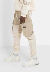 Beige cargobyxor med flera fickor, tapered passform och resårmidja. Bärs med vita sportskor och grå strumpor.
