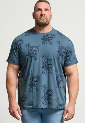 Voksen muskuløs mand med kort hår og skæg iført blå t-shirt med mørke tiger- og bambusmønstre, kombineret med lyseblå jeans.