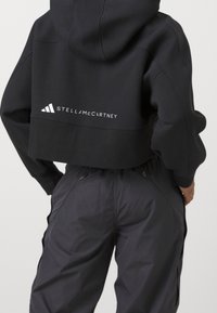 Zkrácená černá mikina s kapucí, která má bílé logo "STELLA McCARTNEY" a tři pruhy na zadní straně, kombinovaná s černými kalhotami.