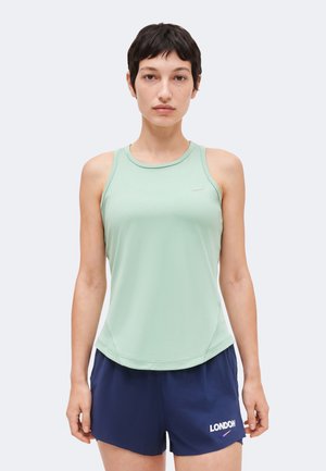 Rövid, sötét hajú nő áll szemben, mentazöld ujjatlan felsőt és sötétkék rövidnadrágot visel, amin a "LONDON" felirat áll.