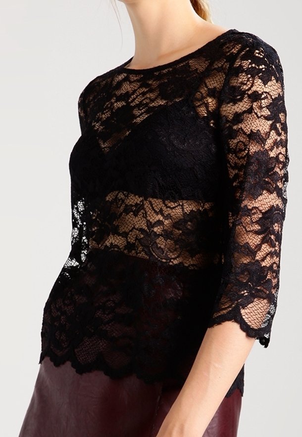 Haut en dentelle noire à manches trois-quarts, avec des motifs floraux et un ourlet festonné. Le tissu est transparent, laissant apparaître une couche inférieure.