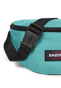 Marsupio teal con una robusta tracolla nera, fibbia in plastica e superficie testurizzata. Include un'etichetta con logo rosso e nero.