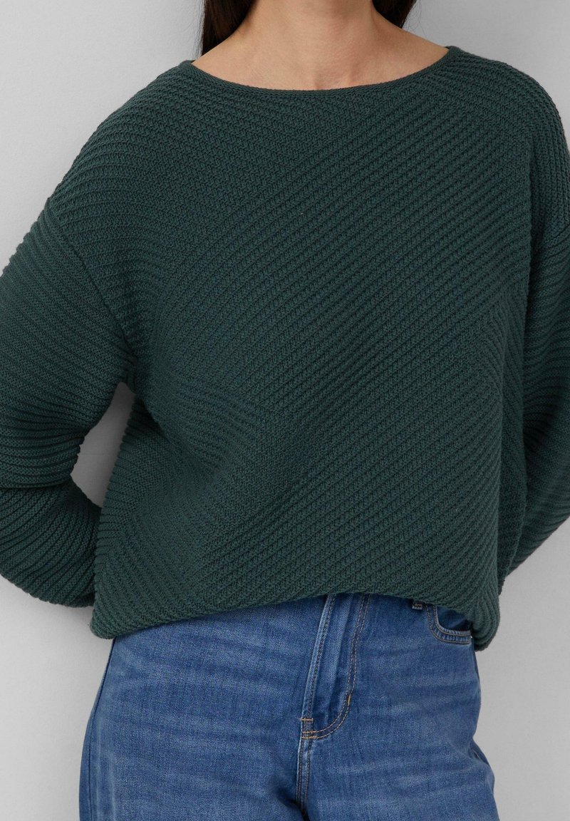 Femme portant un pull en maille texturée vert foncé à manches longues et un jean en denim bleu, debout sur un fond clair uni.
