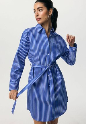 Kobieta poprawiająca pasek sukienki koszulowej w pionowe, niebiesko-białe paski z długim rękawem na jednolitym tle.