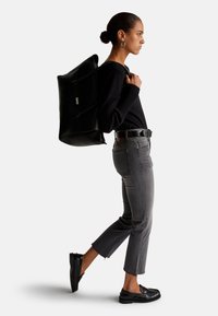 Mochila negra de cuero con un acabado texturizado, llevada sobre un suéter negro y unos pantalones vaqueros grises cortos, combinada con zapatos de charol negros. Diseño sencillo.