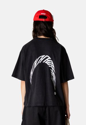 Person med langt mørkt hår bærer en sort oversize T-shirt med hvidt abstrakt bue-design på ryggen og en rød kasket med mærket "WOODBIRD".