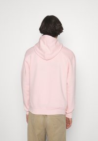 Persona con capelli corti scuri, che indossa un hoodie rosa chiaro e pantaloni beige, di spalle su uno sfondo bianco uniforme.