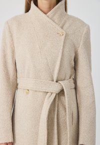 Beige, textúrált anyagból készült átkötős kabát. Jellemzői közé tartozik a sálgallér, a megkötős öv és egy aranyoroszlánfej gomb. Alkalmas hűvösebb időjárásra.