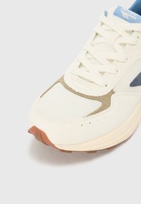 White athletic shoe featuring a mesh upper, beige suede accents, blue logo detail, and a brown rubber outsole with traction patterns.