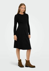 Robe noire côtelée avec des manches longues et un corsage ajusté, évasée à la taille. Portée avec des bottines marron à la cheville avec des accents en métal.