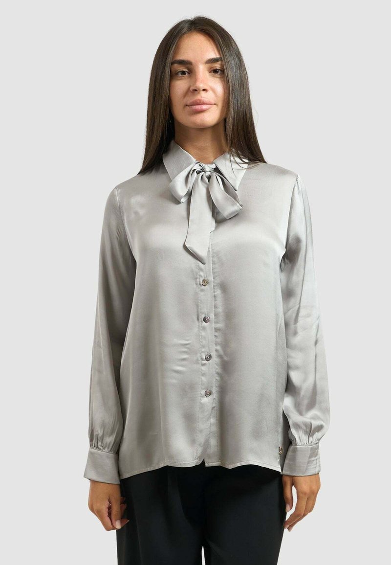 Blusa di seta grigia chiara con colletto a fiocco, maniche lunghe e chiusura con bottoni frontale. Presenta un orlo curvo e dettagli con bottoni sui polsini.
