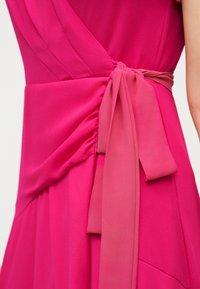 Κοντινή λήψη φωτεινού ροζ φορέματος με ραφές και φαρδιά ζώνη δεμένη σε φιόγκο στη μέση στο πλάι ενός ατόμου.