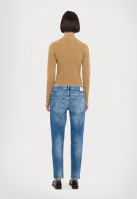Pull en maille côtelée taupe avec col roulé et fermeture éclair au dos, associé à un jean bleu fuselé avec un lavage clair et des poches arrière visibles.